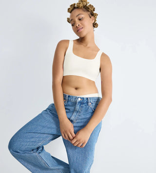 “Modell bär vit Sloggi Zero Feel BH-Top lutat mot en vägg i blåa jeans – osynlig bh-topp med 360° stretch, punktstöd under bysten och sömlös komfort.”